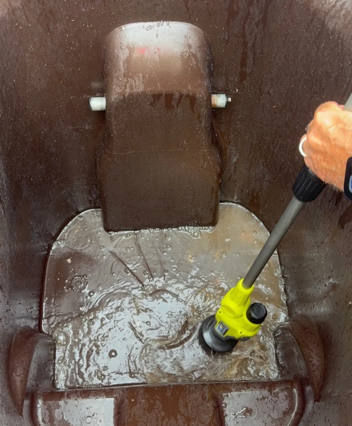 A yellow power scrubber actively cleans the inside of a brown, dirty trash bin filled with murky water.