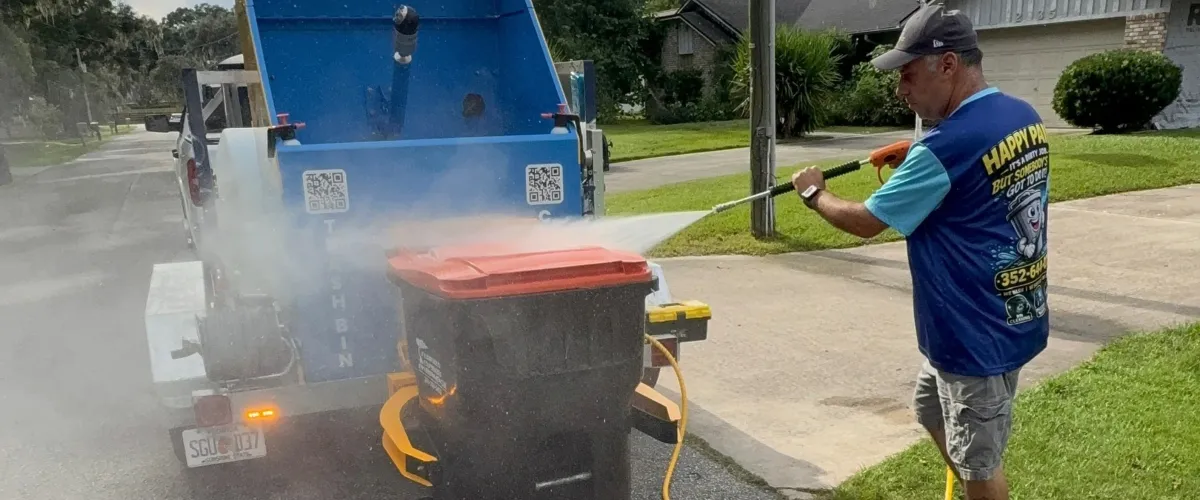 A man in a blue shirt with an American flag patch holds an orange pressure washer gun next to blue cleaning equipment.