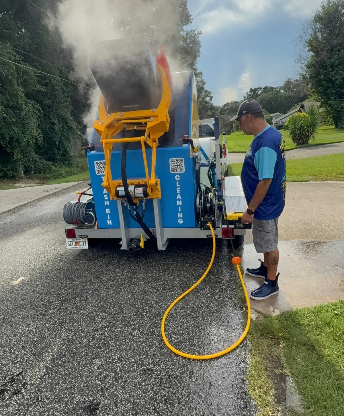 A man in a blue shirt and hat operates a blue and yellow trash bin cleaning machine on a trailer, with steam rising.