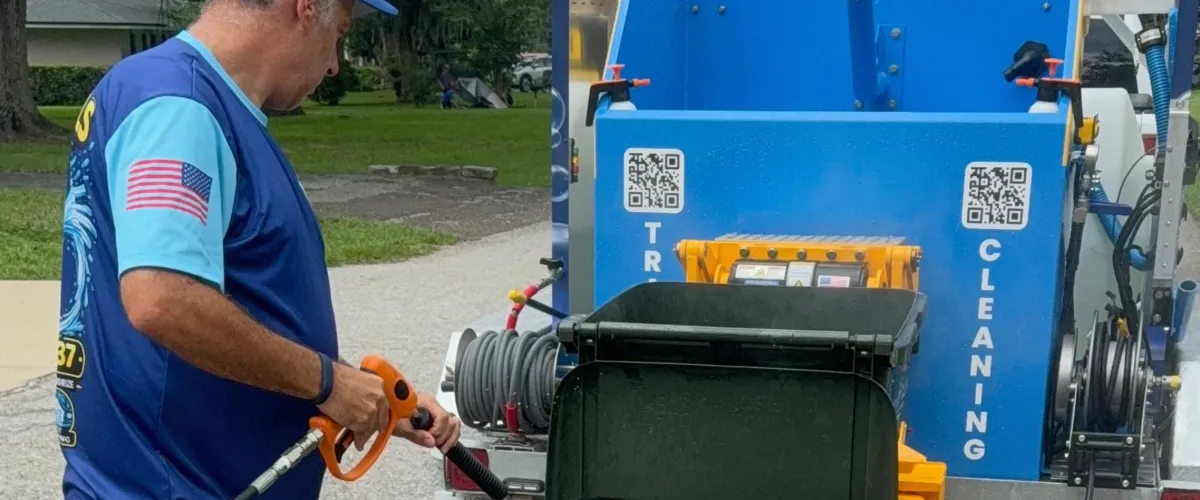 A man in a blue shirt pressure washes a black trash bin mounted on a blue trailer by a residential street.