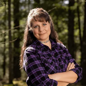 Chris Prebble, LPC, standing in a forest wearing a purple and black flannel shirt, arms crossed, with a confident expression and natural light filtering through the trees.