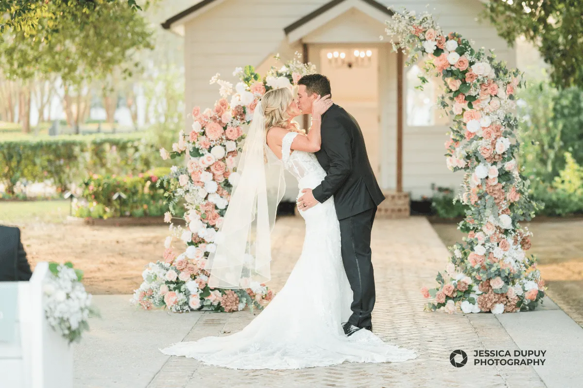 Outdoor wedding ceremony featuring a bride and groom kissing under a lush peach and white floral arch. The setting includes a small white wedding chapel and a stone aisle, captured in a bright and airy photography style.