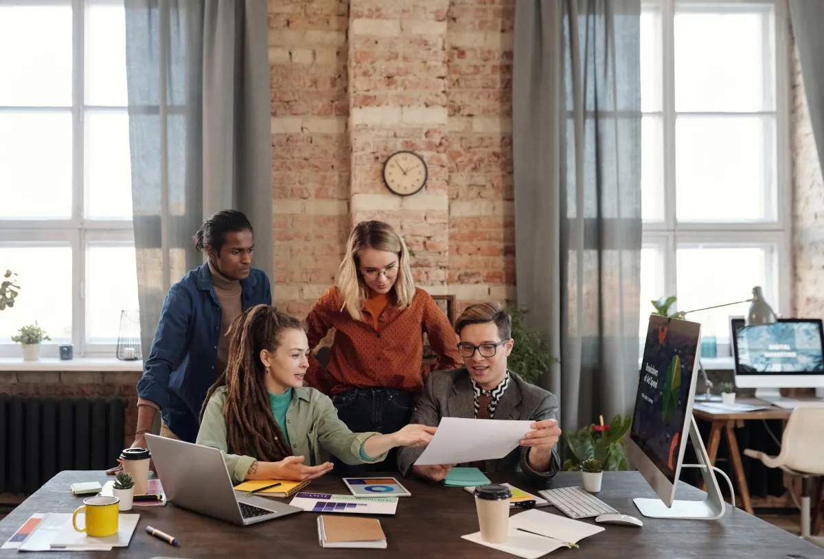 Team of four coworkers reviewing a printed document together around a desk in a modern office.