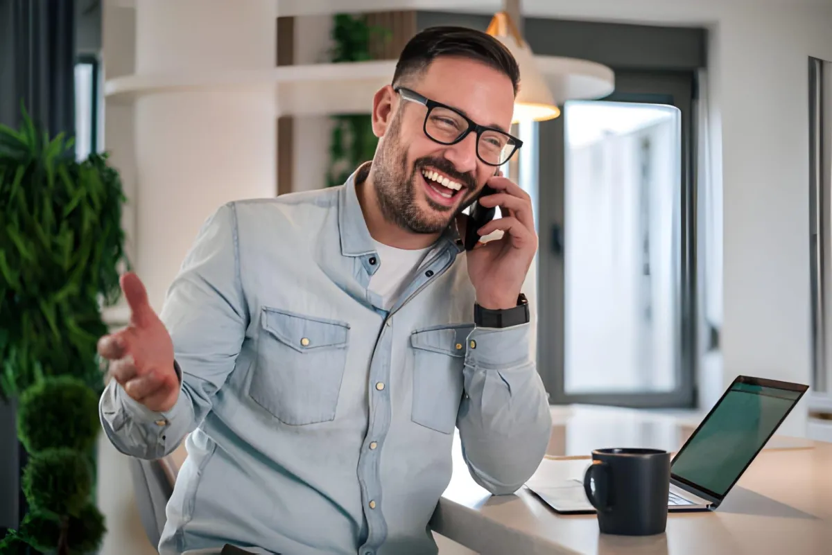 Man in glasses smiling while talking on a phone beside an open laptop and coffee mug in a bright home office.