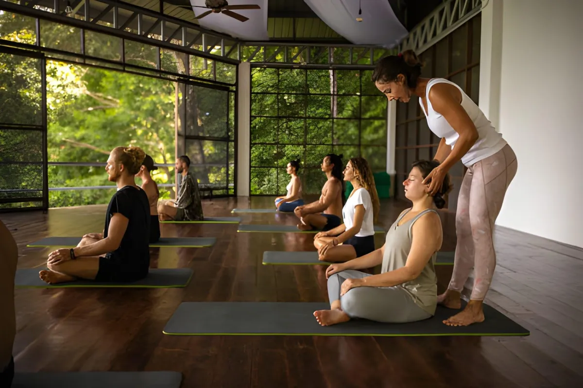 Yoga instructor adjusting a seated student’s posture during a group meditation class in a studio with large windows.