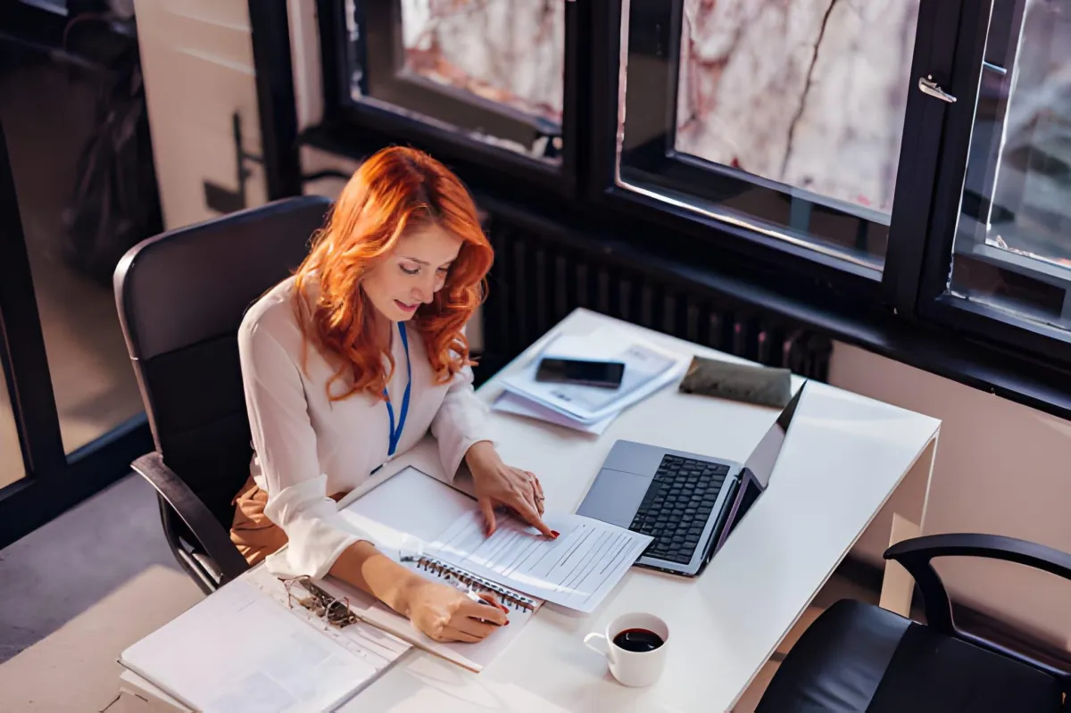 Woman seated at an office desk writing in a notebook beside a laptop, phone, papers, and a cup of coffee