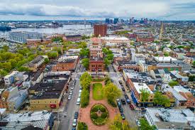 Aerial view of a historic city neighborhood with brick buildings, tree-lined streets, and waterfront skyline in the distance.