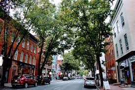 Tree-lined downtown street with historic brick buildings, parked cars, and pedestrians.