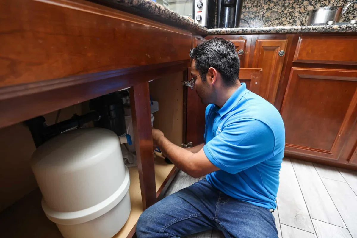 Plumber working under a kitchen sink with a water filtration tank nearby.