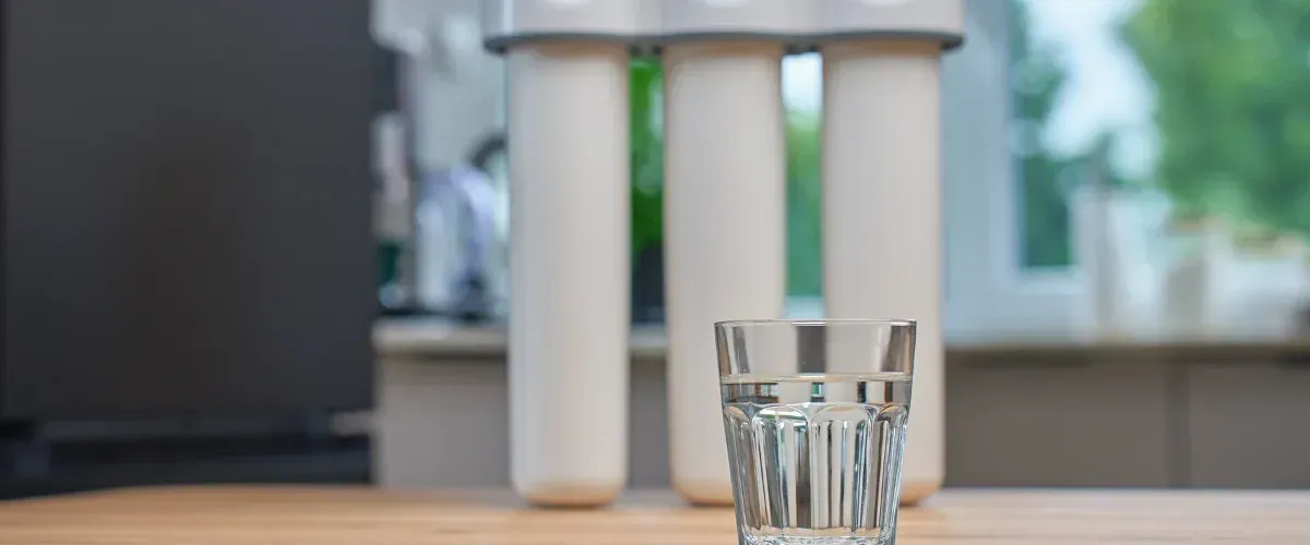 Glass of clean drinking water with home filtration system in background.