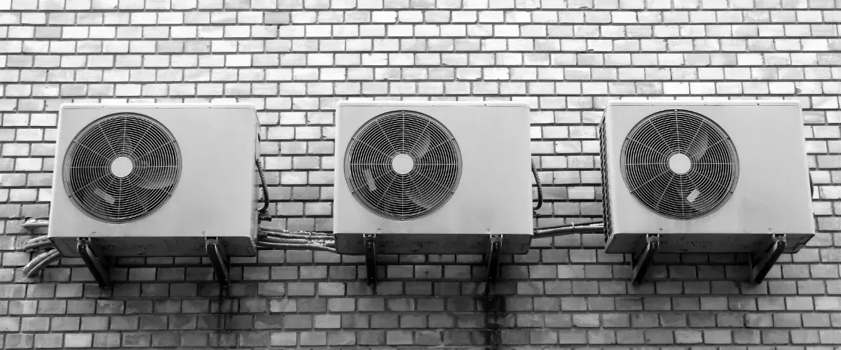 Three outdoor air conditioning units mounted in a row on a brick wall, shown in black and white.