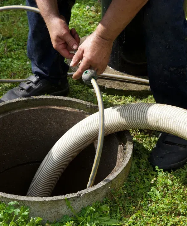 A man's hands handling a hose while standing next to an open septic tank in a grassy area.