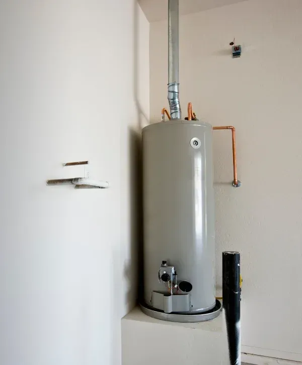 A gray water heater tank with copper and galvanized pipes extending from its top and side, in a corner.