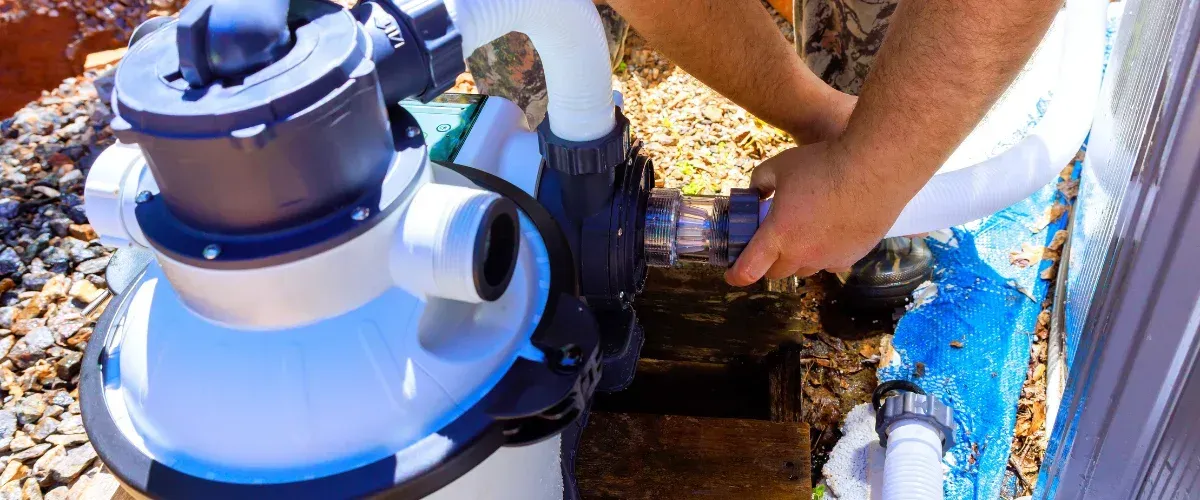 A person connecting a white hose to a pool pump filter system with exposed ground and blue tarp nearby.