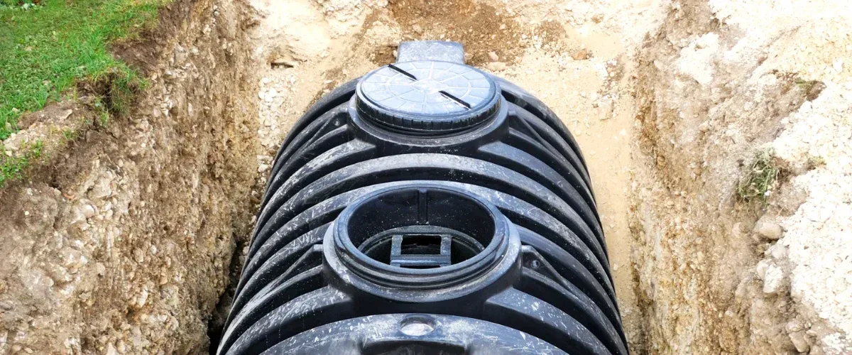 A black corrugated septic tank partially buried in a dirt trench with green grass visible on the left bank.