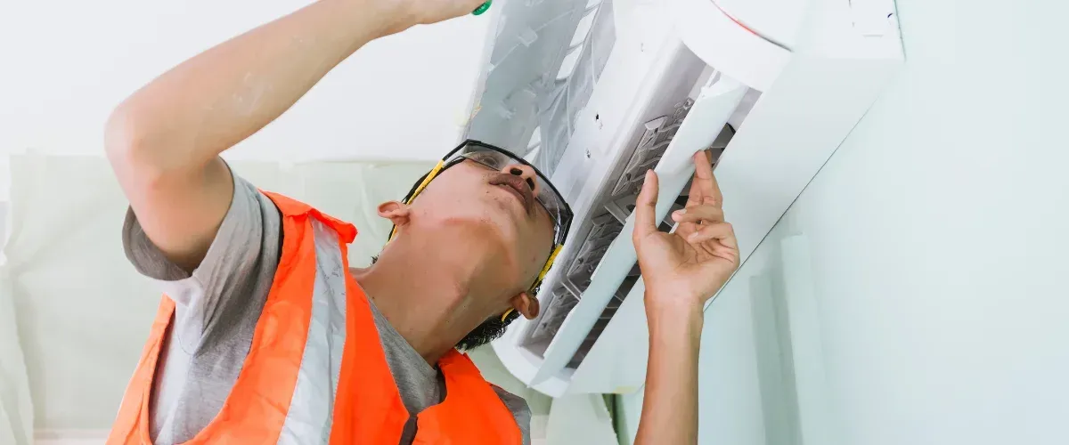A close-up of a technician in a blue uniform adjusting an outdoor air conditioning unit with a tool.