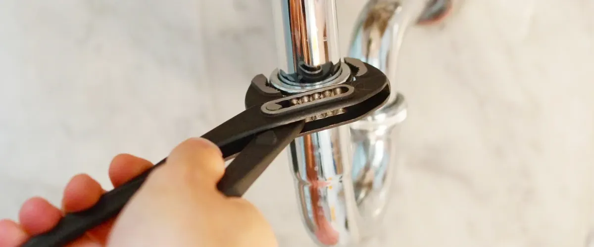 A hand holds a black adjustable wrench, tightening a chrome-plated P-trap pipe under a sink.
