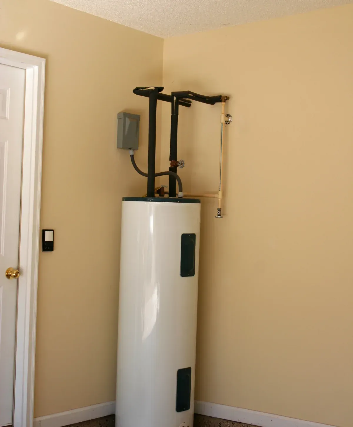 A white electric water heater with pipes and insulation stands in the corner of a room with light beige walls and a white door.