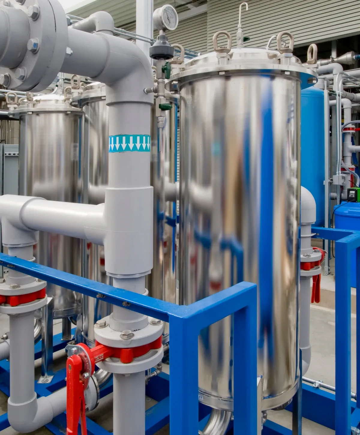 A close-up of industrial filtration equipment with large shiny stainless steel tanks, grey pipes, red valves, and blue metal support frames.