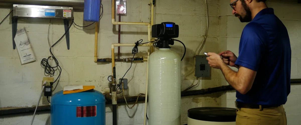 A man in a blue shirt and khakis adjusts settings on a water filtration system in a basement next to pipes and tanks.