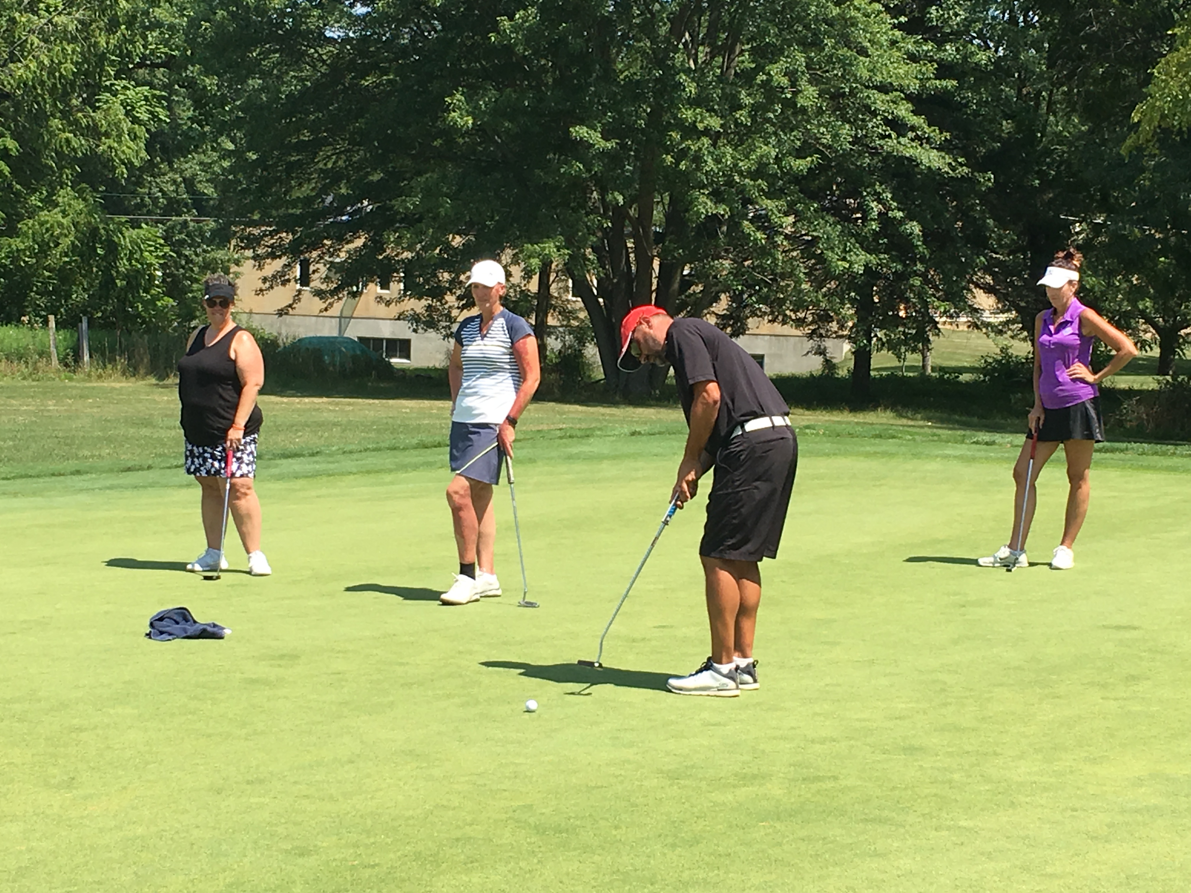 Group of diverse golfers participating in a training session on a practice green, mixed ages and genders, Scott Pieri instructing, sunny afternoon, golf equipment visible, collaborative learning environment, 3:2 aspect ratio.