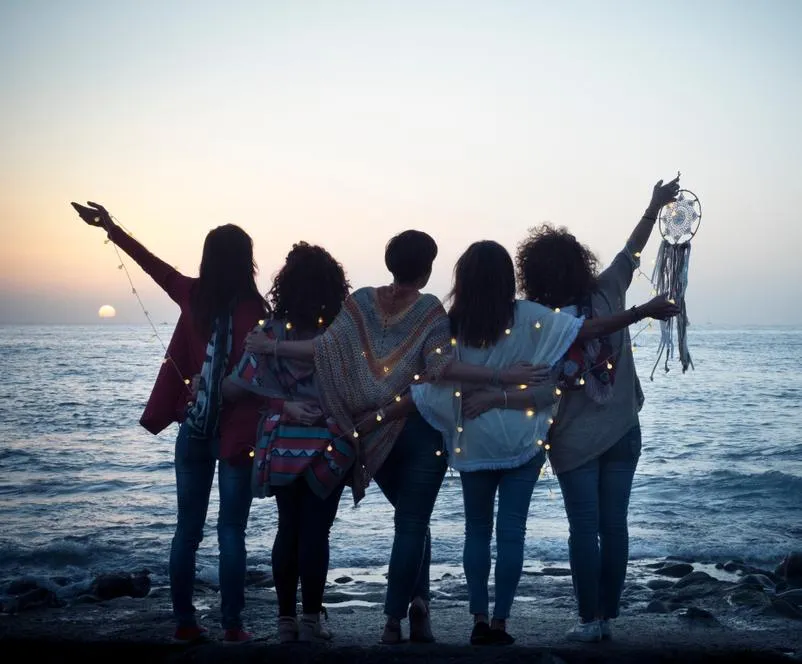 Five women standing at the edge of a body of water, arms entwined and looking out at the setting sun.