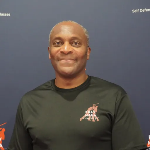 Female instructor, mid-30s, demonstrating a defensive block, short hair, athletic build, smiling, in a bright training room, 1:1 aspect ratio
