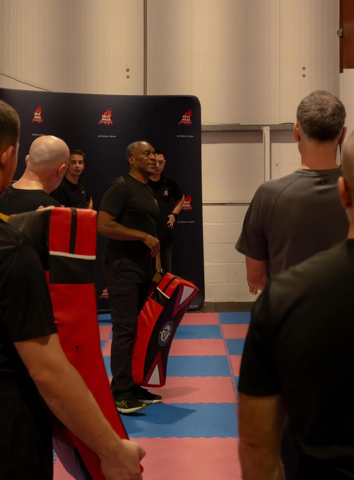 Male Krav Maga instructor demonstrating a self defence move with a smiling participant, both mid-action, in a well-lit gym, diverse adults