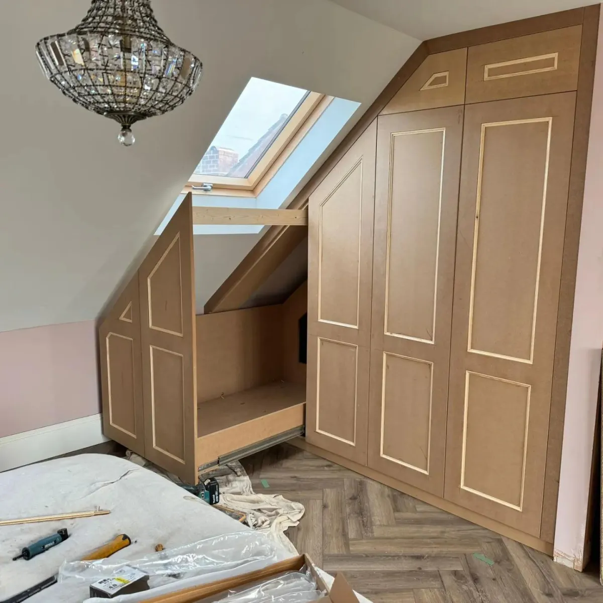 Custom-designed angled fitted wardrobe in an under-eaves space, crafted by a skilled carpenter in the West Midlands, UK, featuring elegant panel doors, bespoke storage solutions, and a neat finish that complements the room's sloped ceiling and hardwood flooring, mid-installation with tools visible.