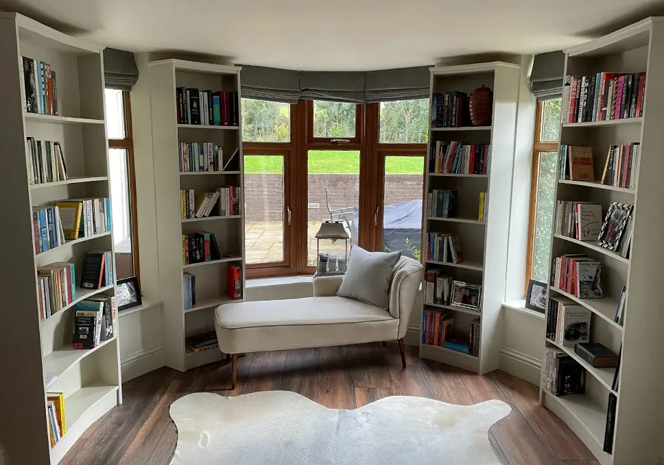Luxurious home library with custom-built, white bookcases framing a bay window, expertly designed by a West Midlands carpenter for a personalized touch to the home, featuring a comfortable chaise lounge and plush rug, with a serene garden view, ideal for a peaceful reading nook in the UK.
