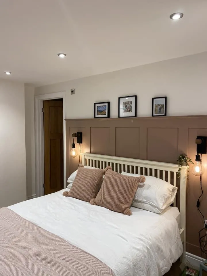 Elegant bedroom featuring a custom-made, panelled headboard in a warm taupe shade by a West Midlands carpenter, with coordinated bedding and pillows, stylish wall-mounted bedside lamps, framed pictures above the bed, and a sophisticated ambiance enhanced by recessed ceiling lighting.