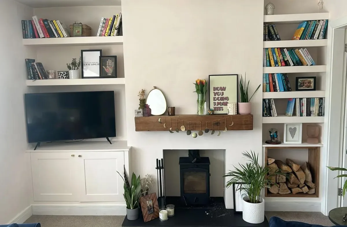 Bespoke white fitted alcove units with shelves and cabinets flanking a black wood burner fireplace, handcrafted by a West Midlands-based carpenter and joiner, Edwoodwork, showcasing custom shelving filled with books, personal mementos, and indoor plants, enhancing a cosy living room interior.