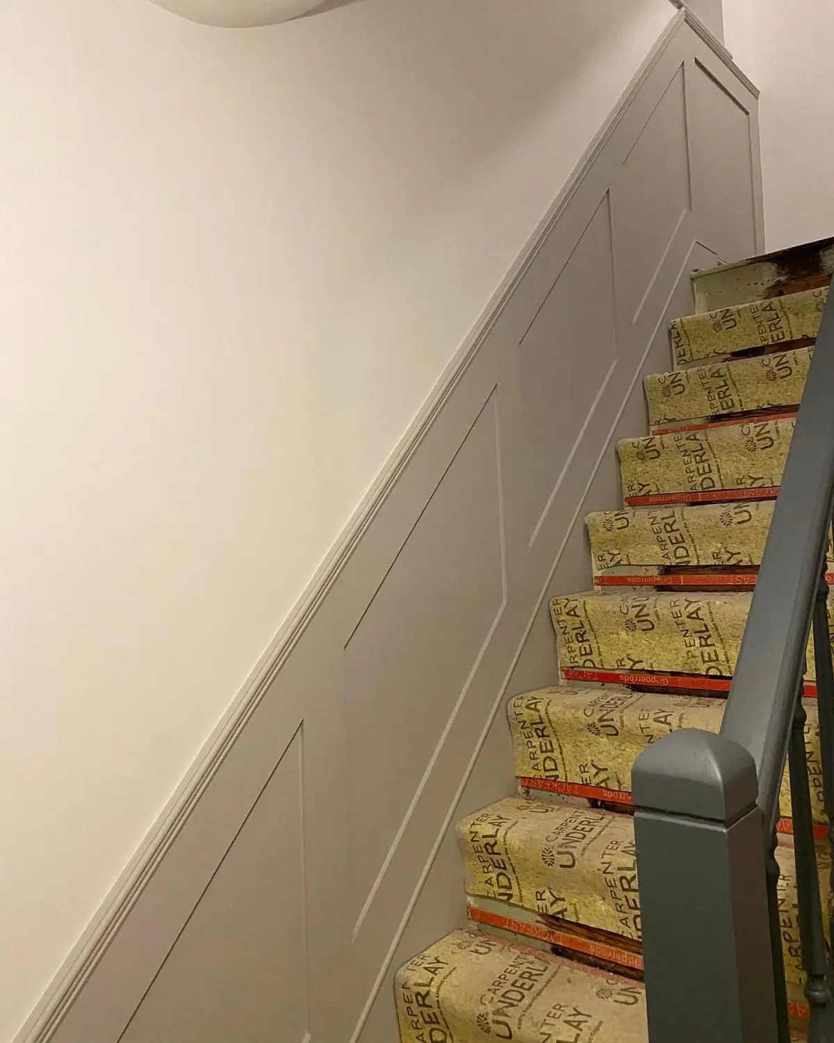 A staircase with sophisticated wall paneling in a soft grey, contrasting with the natural wood of the steps and handrail, all custom-fitted by a skilled West Midlands joiner, showcasing a blend of classic design and contemporary craftsmanship in a home undergoing stylish renovations.