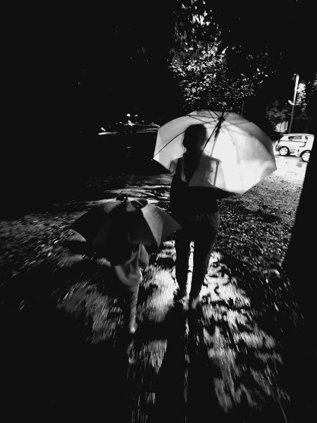 Adulto e bambino camminano sotto la pioggia con un ombrello, immersi in un’atmosfera scura e sfocata che richiama vulnerabilità, protezione e attraversamento condiviso di un momento difficile.