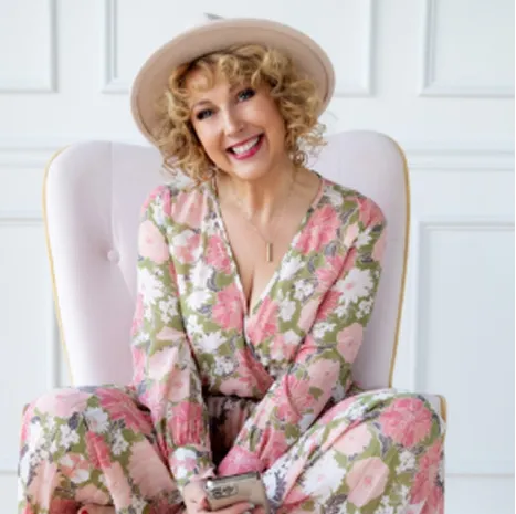 Woman sitting in a white chair, wearing a floral jumpsuit and wide-brim hat, smiling warmly
