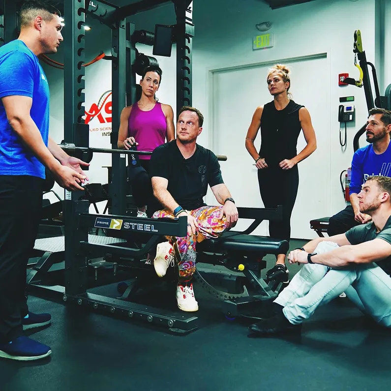 Small group of individuals engaged in a fitness and coaching session at a gym, reflecting teamwork, mentorship, and lifestyle alignment.