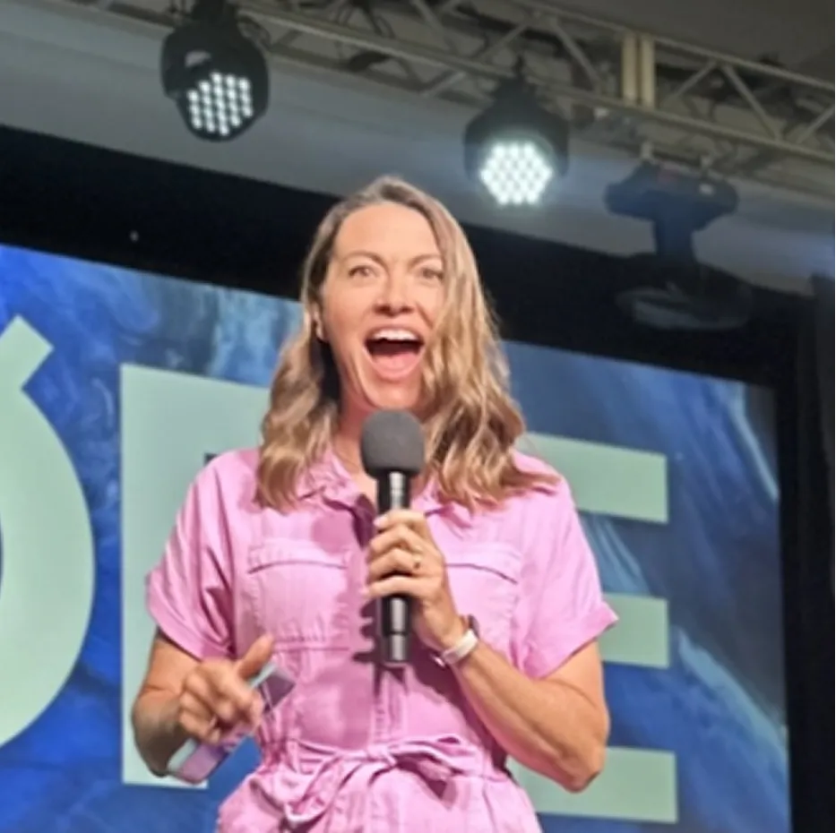 Woman in a pink jumpsuit speaking on stage with a microphone, mid-sentence, expressive facial expression