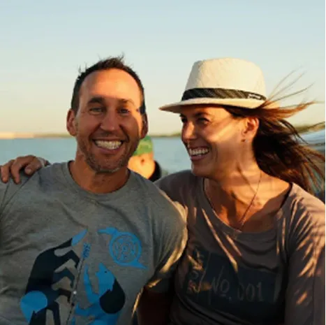 Man and woman smiling near a body of water during golden hour; woman wearing a sun hat