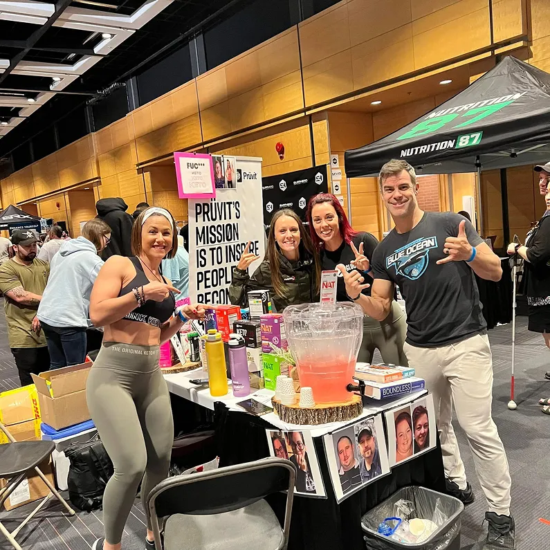 Entrepreneurs smiling at a booth showcasing health and wellness drinks at a busy expo, highlighting community outreach and brand promotion.