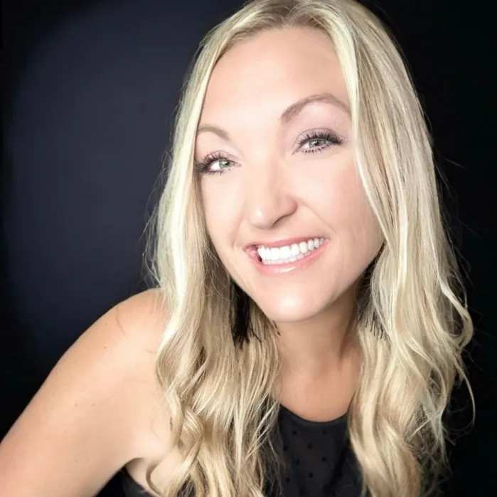 Woman with long blonde hair wearing a light denim shirt, smiling with natural light behind her