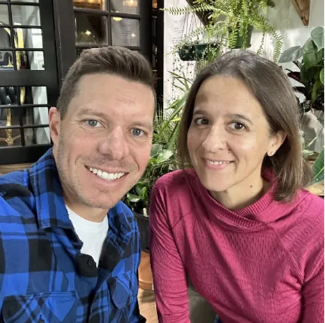 Man in a blue plaid shirt and woman in a pink sweater smiling together indoors with plants in the background