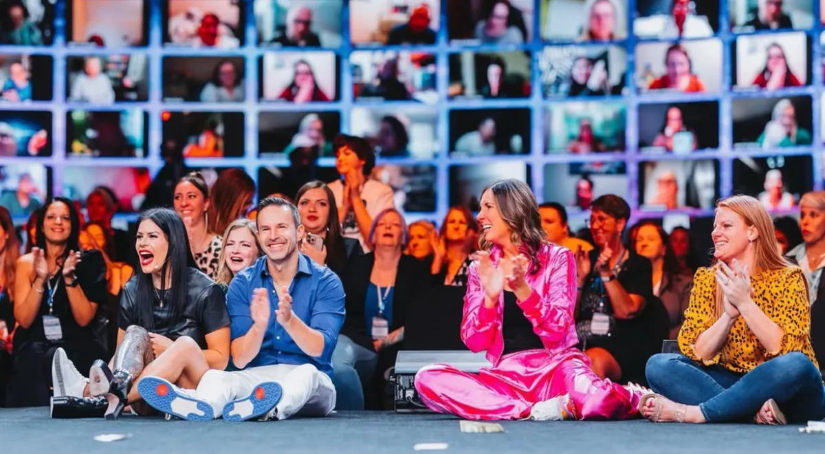 A diverse group of excited attendees clapping and smiling at a live event, with a vibrant audience and digital wall of virtual participants in the background, showcasing community engagement and team spirit at Momentum Pro.