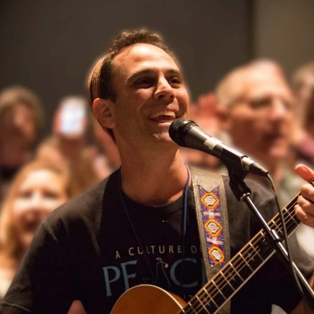 Craig Parks performing with a guitar and microphone, smiling on stage in front of a blurred audience, wearing a shirt that reads “A Culture of Peace.”