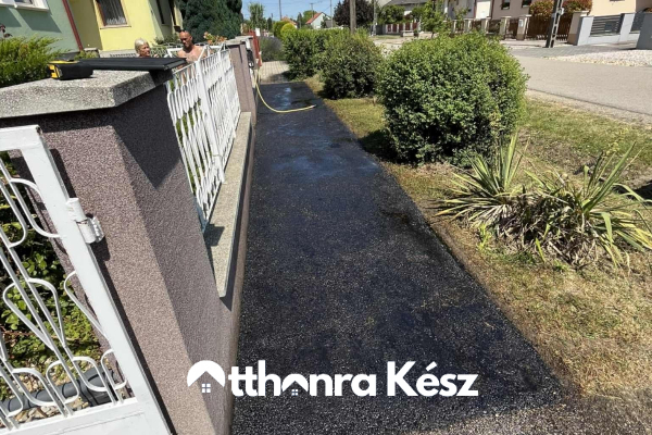 Keskeny, frissen aszfaltozott bejáró kerítés és kertszegély mellett, rendezett fekete aszfalt felülettel