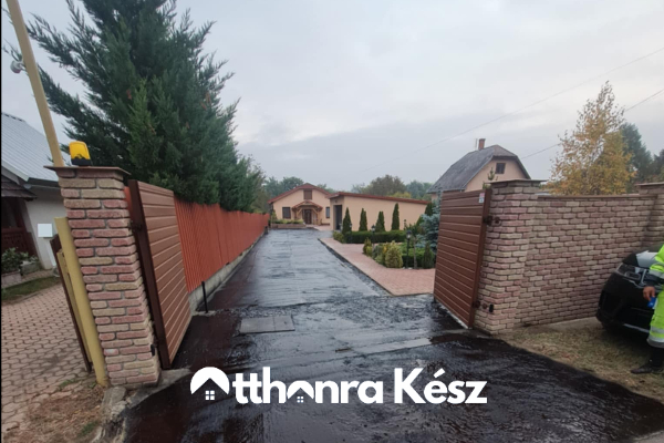 Frissen aszfaltozott kapubejáró nedves felületen, kétoldali kerítéssel és rendezett fekete aszfalt burkolattal