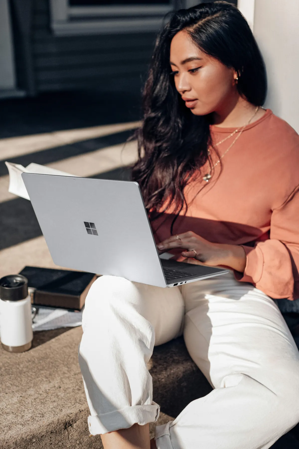 A brown-skinned young woman with dark hair, orange shirt, and white pants sits on a concrete step.  Her laptop is open on her lap, a book and water bottle are beside her.