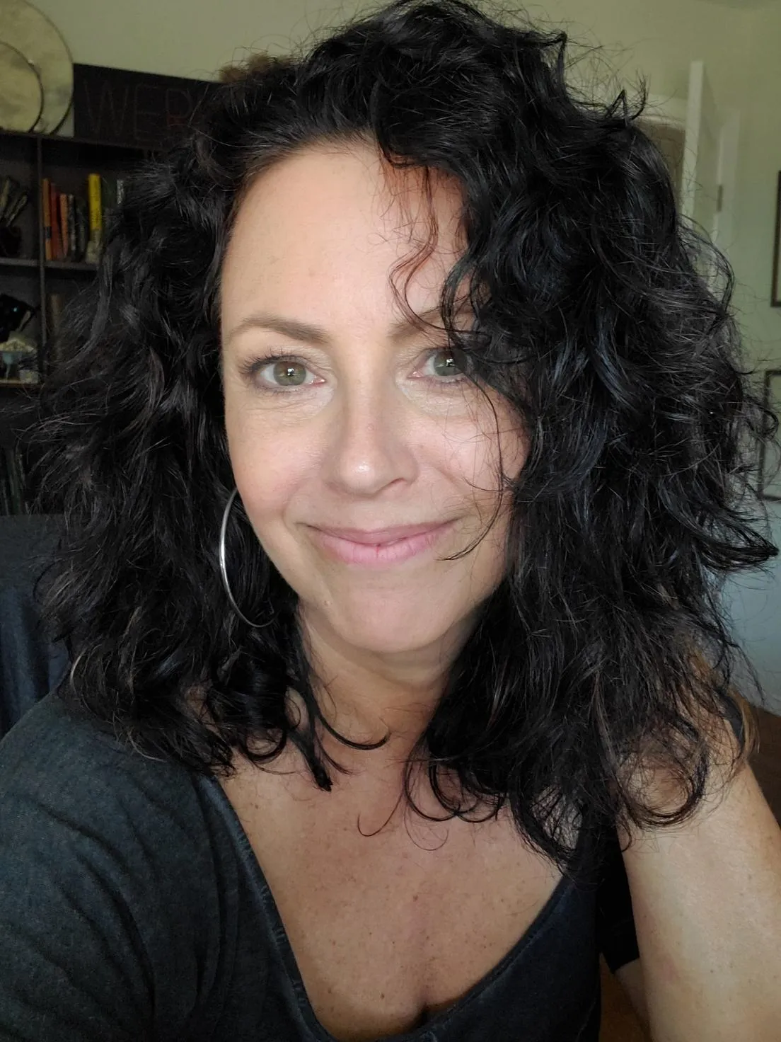 A middle-aged white woman with dark, wavy hair and silver hoop earrings is smiling at you