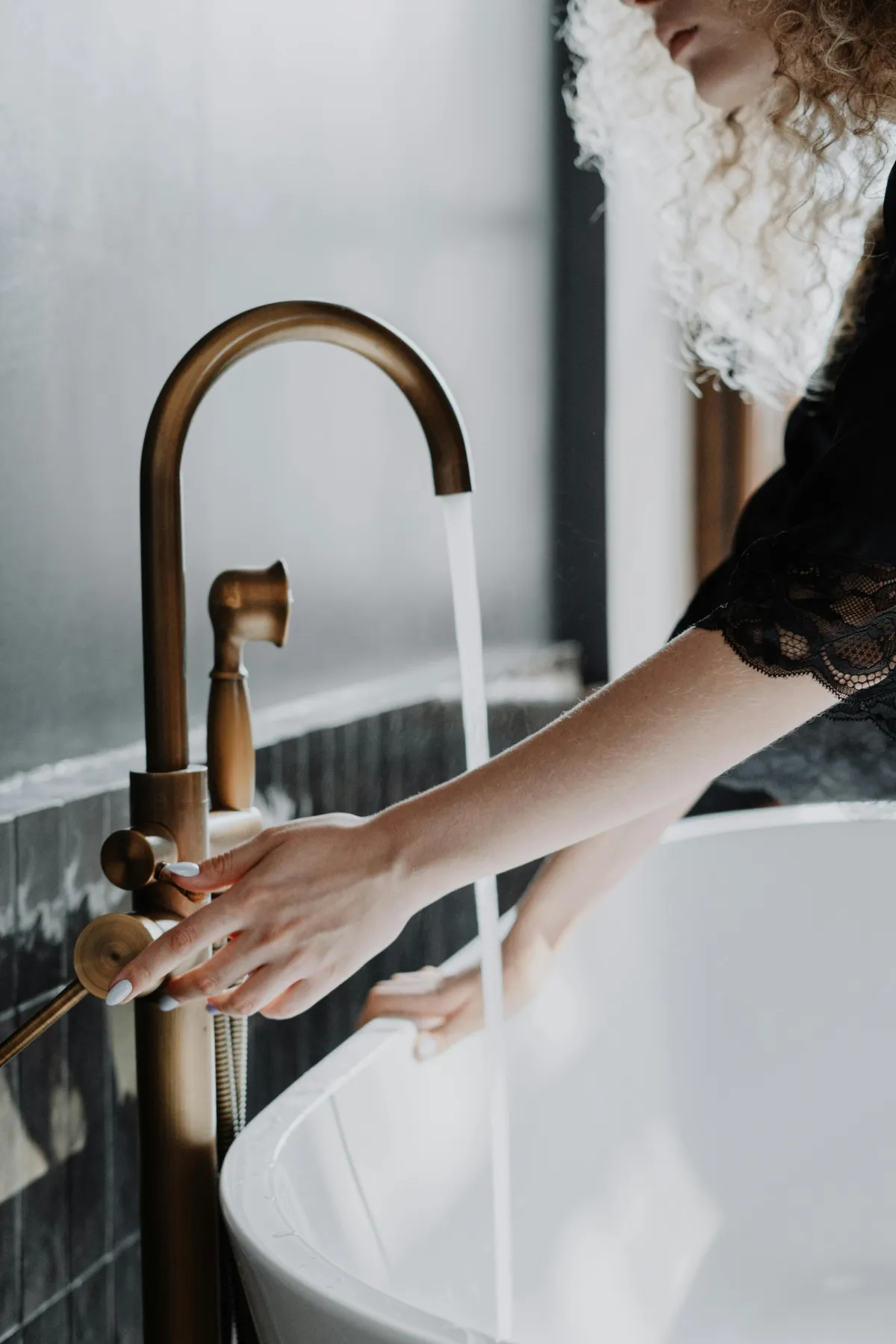 Woman adjusting a modern brass faucet while filling a white bathtub, illustrating plumbing services for commercial properties.
