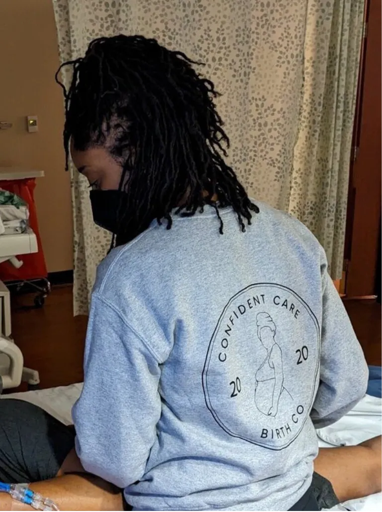 Birth worker wearing a ‘Confident Care Birth Co.’ sweatshirt providing hands-on support to a laboring woman