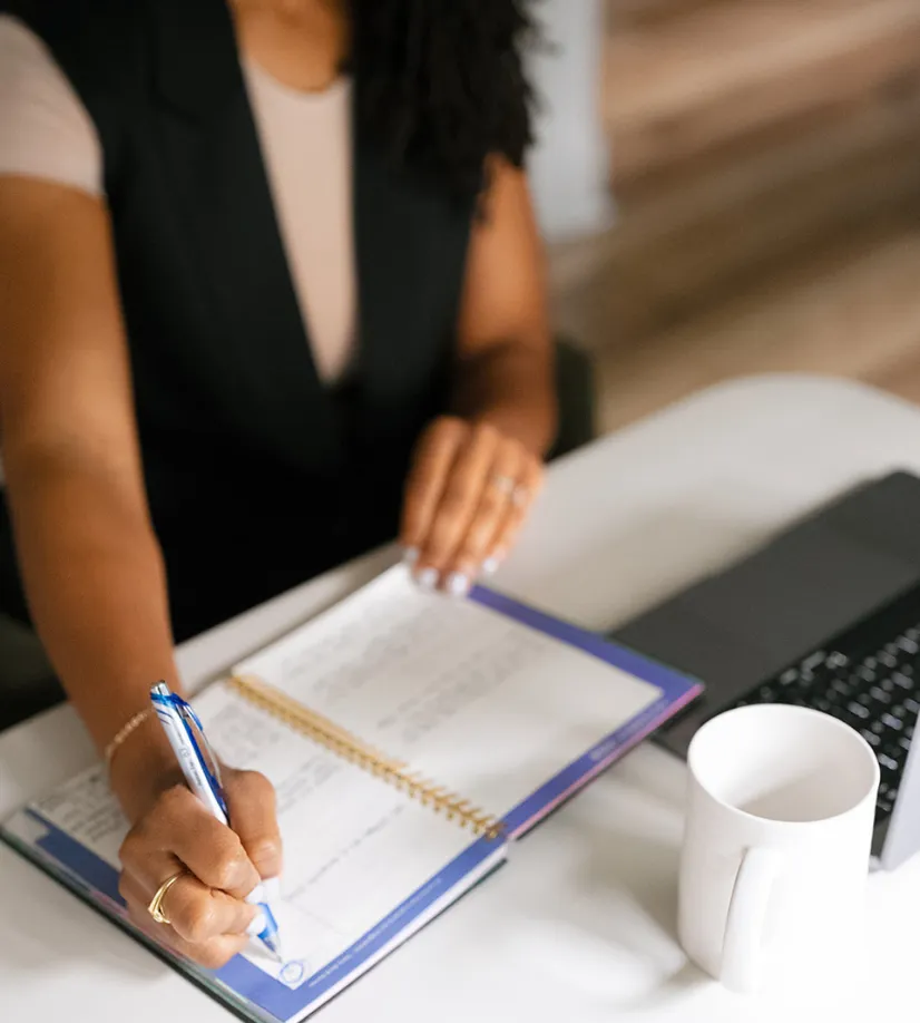 Woman writing in a planner next to a laptop and coffee mug